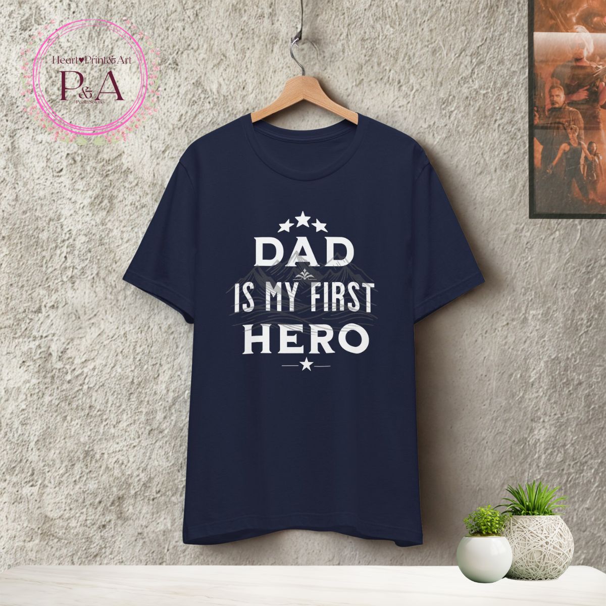 Navy 'Dad Is My First Hero' t-shirt on hanger with wooden chair and white cap.