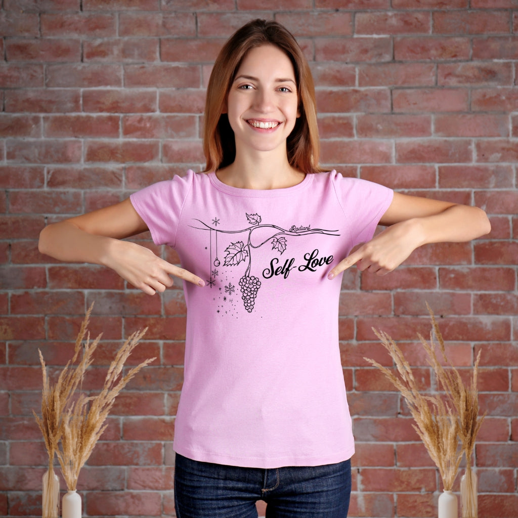 A woman wearing a pink t-shirt featuring the words "self-love" and a grapevine design, promoting self-love.